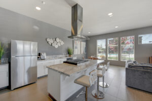 Interior Clubhouse, Open Floor plan Kitchen, Large island with breakfast bar and 3 seats, circular 4 seat table right of kitchen island, stainless steel appliances, marble like countertops, contemporary art and design throughout room, Gray Walls.