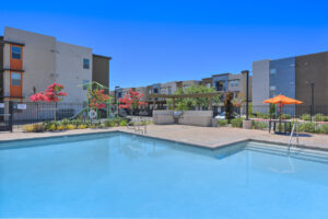 Exterior Swimming Pool, Grill area in corner of enclosed pool area, bistro tables, playground outside of fence, residential buildings in background.